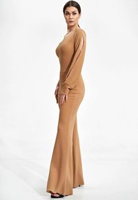 Bruine jumpsuit met lange mouwen, uitlopende pijpen, uitsparingen bij de schouders, gemaakt van soepel materiaal. Gedragen met zwarte hoge hakken sandalen.