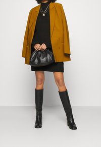 Senapsgul kavaj, svart stickad klänning, svarta läderstövlar till knäna och en svart väska. Lager av halsband ger subtila detaljer.