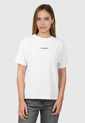 Giovane donna con capelli castani lisci che indossa una T-shirt bianca con piccole scritte nere "LIEBE AUF DIE EINS" e jeans grigio scuro, su sfondo grigio chiaro.