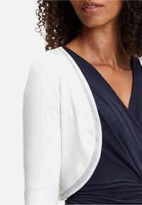 Chaqueta blanca de corte abierto con textura suave, con un vestido envolvente de color azul marino debajo que contrasta. Detalle sutil de ribete gris a lo largo del borde de la chaqueta.