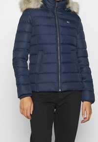 Frau trägt eine marineblaue gesteppte Daunenjacke mit Kapuze aus Fell, einem Reißverschluss auf der Vorderseite und Seitentaschen, kombiniert mit schwarzen Hosen.
