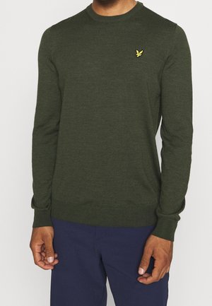 Mand iført mørkegrøn crewneck-trøje med lille gul fuglelogo på brystet og mørkeblå bukser, stående med armene afslappet.