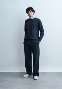 Moški model z zavitega lasmi, oblečen v mornarsko modro majico z dolgimi rokavi in ujemajoče široke hlače, bele superge, stoji z rokami v žepih pred enobarvnim ozadjem.