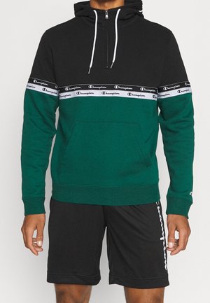 Mężczyzna w czarno-zielonej bluzie z kapturem Champion z białymi paskami z logo na piersi i rękawach, w połączeniu z czarnymi szortami Champion.
