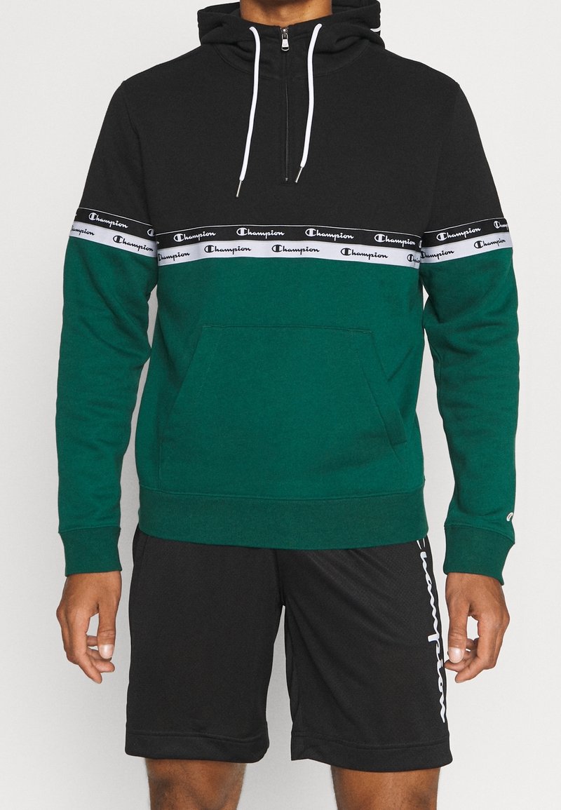 Homme portant un sweat à capuche Champion noir et vert avec des bandes blanches avec le logo sur la poitrine et les manches, associé à un short noir Champion.