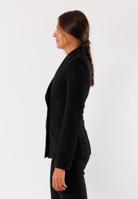 Blazer nero su misura con spalle strutturate, chiusura a un bottone e vestibilità slim. Tessuto liscio e tasche laterali. Indossato con pantaloni abbinati.