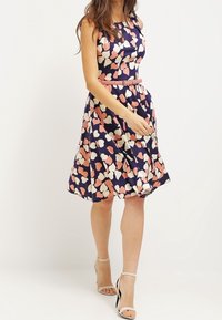 Ärmelloses Kleid mit Blumenmuster in Marineblau, das rosa und cremefarbene Akzente aufweist, eine taillierte Taille mit einem passenden Gürtel hat und einen weit auslaufenden Rock besitzt.