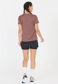 Kvinna med flätat hår som bär en mauve kortärmad tröja, svarta shorts, vita strumpor och silvriga löparskor, stående mot en vit bakgrund.