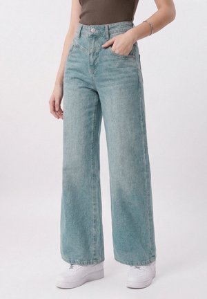 Person som har på seg lyseblå jeans med høyt liv og vide ben, hvite sneakers, brunt ermeløst topp og et sølvarmbånd på høyre håndledd.