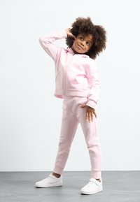 Uma sweatshirt de fecho éclair rosa claro e calças de treino combinando, feitas de tecido macio. Ténis brancos completam o visual. A criança posa com uma expressão brincalhona.
