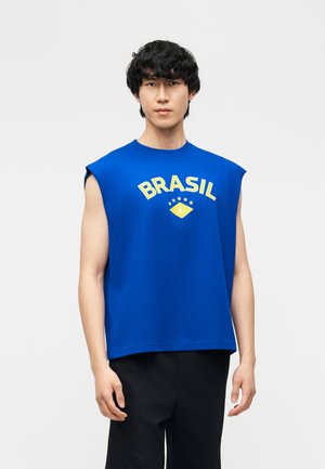 Jeune homme aux cheveux noirs portant un débardeur bleu avec l’inscription « BRASIL » en jaune, associé à un pantalon noir, debout devant un fond uni.