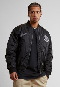 Jachetă bomber neagră din nylon lucios, cu manșete și tiv cu nervuri. Prezintă logo-ul brodat "Starter" și fermoar pe lateral.