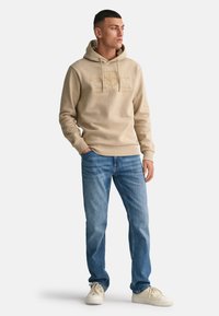 Felpa beige con cappuccio, tasca a marsupio, coulisse e logo in rilievo, abbinata a jeans azzurri dritti e sneakers bianche.