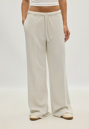 Personne portant un pantalon ample beige clair avec cordon de serrage et coutures avant, ainsi que des chaussures blanches à enfiler, debout les mains dans les poches.
