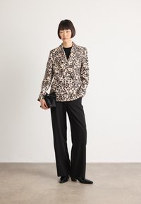 Vrouw staat in een blazer met luipaardprint, zwarte wijde pantalons, zwarte laarzen, en houdt een kleine zwarte clutch vast tegen een effen achtergrond.