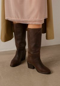 Botas de cuero marrón hasta la rodilla con un tacón cuadrado, que presentan una textura suave y una puntera redondeada, combinadas con una falda rosa claro y un abrigo verde.