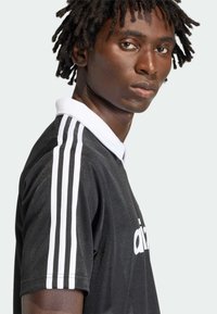 Μαύρο πόλο πουκάμισο με λευκές λεπτομέρειες και τρεις ρίγες στα μανίκια, με γιακά και αθλητική εφαρμογή. Το υλικό φαίνεται λείο και με υφή.