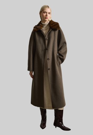 Long manteau en laine marron avec un col en fourrure, doté d'une fermeture à boutons et de poches latérales, porté par-dessus une robe claire et des bottes marron.