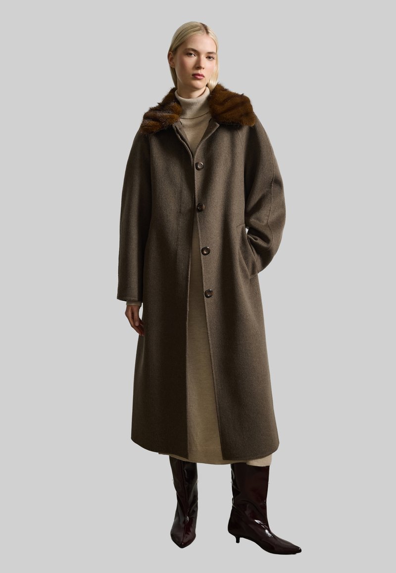 Cappotto lungo in lana marrone con colletto in pelliccia, dotato di chiusura con bottoni e tasche laterali, indossato sopra un vestito chiaro e stivali marroni.
