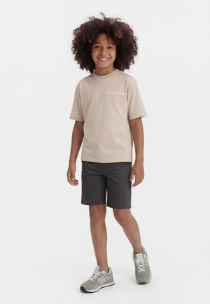 Bambino con capelli ricci che indossa una maglietta beige New Balance, pantaloncini scuri e scarpe da ginnastica grigie New Balance, in piedi e sorridente.
