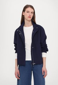 Navy blue lightweight jacket with a zip closure, snap buttons, and elastic cuffs, layered over a white shirt, paired with blue jeans.