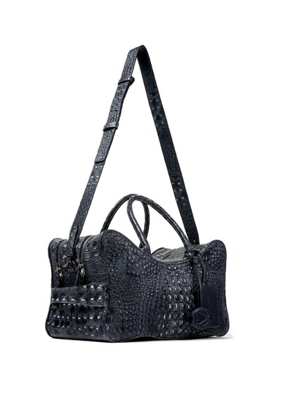 Sac à main en cuir noir texturé avec deux poignées, bandoulière ajustable et étiquette de bagage attachée, suspendu sur un fond blanc.