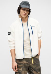 Νέος άνδρας που φοράει λευκή φλις ζακέτα με μπλε περιθώριο, μπεζ πουκάμισο, παντελόνι παραλλαγής τύπου cargo και σκούφο σκούρου γκρι χρώματος, στέκεται μπροστά σε απλό φόντο.