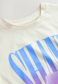 T-shirt en coton couleur crème avec un col rond et un grand texte dégradé bleu-violet partiellement visible.