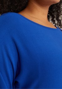 Blue knit top featuring a wide neckline and ribbed trim, with a smooth texture and relaxed fit. Close-up view emphasizes color and material.