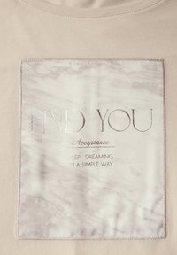 Světle béžové tričko s čtvercovou grafikou s mramorovým vzorem, stříbrným nápisem "FIND YOU Acceptance" a dekorativním šitím.