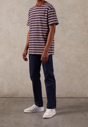Homme portant un T-shirt à rayures courtes bleu marine, blanc et marron, un jean bleu foncé et des baskets blanches, debout sur un sol en béton.