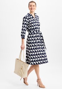 Marineblaues Kleid mit einem weißen Wellenmuster, langen Ärmeln, einem Gürtel in der Taille und knielanger Schnittführung. Begleitet von einer beigefarbenen Tote-Tasche.