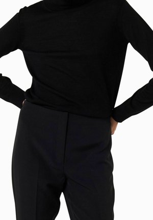 Personne portant un haut noir à manches longues et un pantalon taille haute noir, les mains posées sur les hanches sur un fond blanc uni.