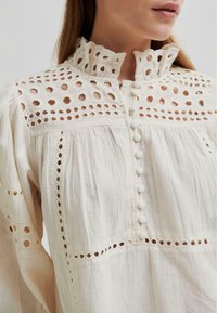 Weiße Baumwollbluse mit hohem Kragen und Rüschendetails, dekorativen Ausschnitten und Stoffknöpfen entlang der Frontleiste.