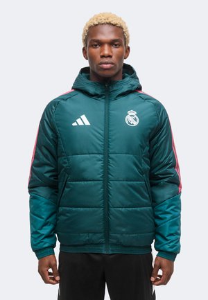 Man met kort blond haar die een blauwgroene Adidas Real Madrid gewatteerde jas met roze strepen draagt, staand tegen een effen achtergrond.