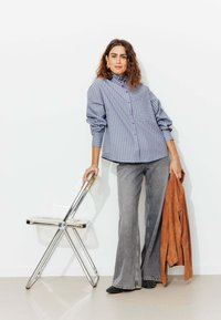 Chemise boutonnée à rayures bleu et blanche avec un col et une poche poitrine, associée à un jean évasé gris, tenant une veste marron et une chaise métallique.