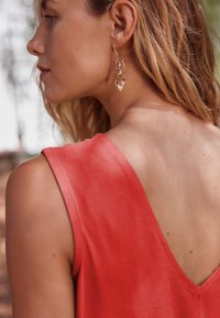 Haut sans manches corail avec un col en V, texture lisse. Boucles d'oreilles dorées avec des perles transparentes et des accents suspendus au lobe de l'oreille.