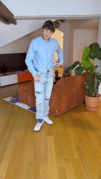 Camicia azzurro chiaro, jeans azzurro chiaro strappati, sneakers bianche, in piedi in una stanza con pavimento in legno, un divano in corduroy marrone e una pianta.
