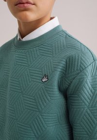 Zelená textúrovaná mikina so šesťuholníkovým vzorom, rebrovaným golierom a vyšívaným logom na hrudi. Biela košeľa s golierom pod mikinou.