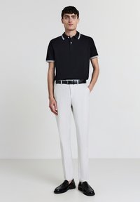 Schwarzes Polohemd mit weißen Akzenten, kombiniert mit hellgrauen Hosen. Das Modell trägt polierte schwarze Schuhe, verfügt über kurze Ärmel und hat ein tailliertes Design.