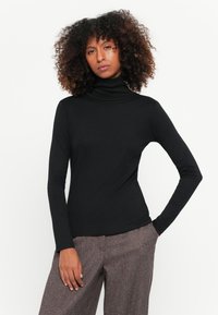 Schwarzer Rollkragenpullover mit figurschmeichelndem Design und langen Ärmeln. Glattes, dehnbares Material. Das Modell trägt ihn mit hoch taillierten Hosen.