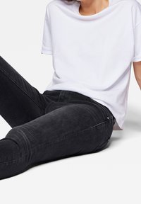 Camiseta blanca de algodón con mangas enrolladas, combinada con jeans oscuros de color gris que tienen un ligero estiramiento y un desvanecimiento sutil, apoyada contra un fondo claro.
