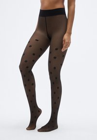 Collants noirs à pois avec une texture transparente, dotés d'une large ceinture haute noire unie et de grands motifs circulaires sur un fond sombre.