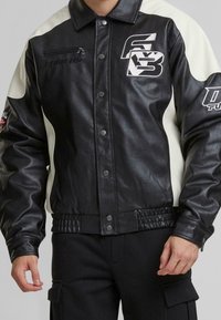 Veste en cuir noir et blanc avec une fermeture à pression, une ceinture côtelée et une poche zippée sur le côté ; comporte des détails de logo sur la poitrine et la manche.