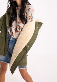 Zelená parka s pohodlnou béžovou umělou kožešinovou podšívkou, vybavená druky a stahovací šňůrkou, doplněná světlým vzorovaným topem a džínovými šortkami.