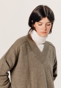Olivegrüner Wollpullover mit V-Ausschnitt und überschnittenen Schultern, über einem weißen gerippten Rollkragenpullover getragen. Weiche Textur, minimalistische Details.