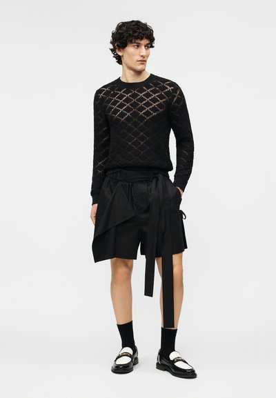 Άνδρας με κατσαρά μαλλιά που φοράει μαύρο πουλόβερ ανοιχτής πλέξης, ασύμμετρα μαύρα σορτς με κρεμαστές λωρίδες υφάσματος, μαύρες κάλτσες και ασπρόμαυρα μοκασίνια.