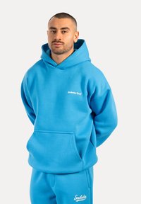 Sweatshirt à capuche bleue en tissu doux, avec une poche kangourou et un logo sur la poitrine. Associé à un pantalon de survêtement bleu assorti.