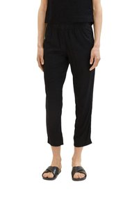 Persoon in zwarte cropped broek met elastische taille, gecombineerd met zwarte schuifsandalen, staand tegen een witte achtergrond.