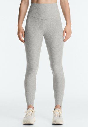 Kobieta ubrana w jasnoszare legginsy z wysokim stanem i szczegółami z paskami nad beżowymi sneakersami, stojąca na jednolitym białym tle.